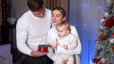 beautiful family of three standing near the christmas tree. parents make a wish and blow the candle on the little cake in man's hand. vertical video.