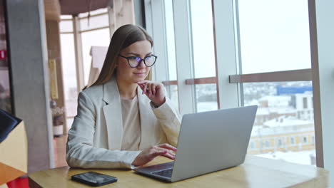 positive young female working in the light office. attractive long-haired lady wearing glasses surfing internet on her laptop.