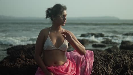 woman in white bikini and pink sarong sitting on rocks by the sea, wind in hair, contemplative mood, daylight