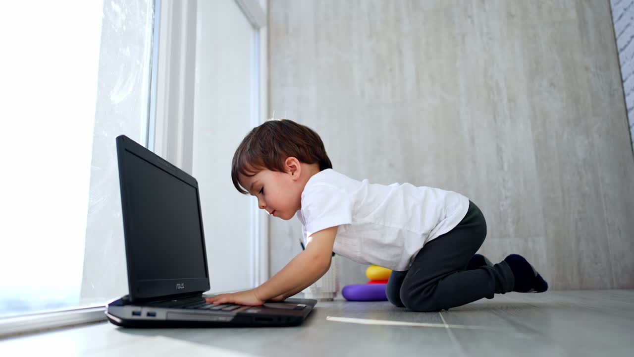 Cute baby boy playing with an old laptop. Pretty toddler presses keys and closes the gadget. Low angle view.
