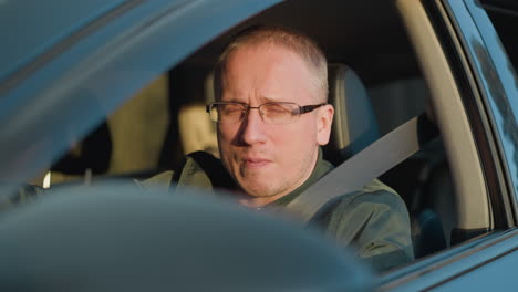 close-up front view of a man wearing glasses and a green jacket sitting in the driver s seat of a car. he is looking straight ahead with a serious expression, exuding focus and determination