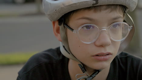 cute teen kid with glasses and a bike helmet sitting outside looks down and then looks up and off camera