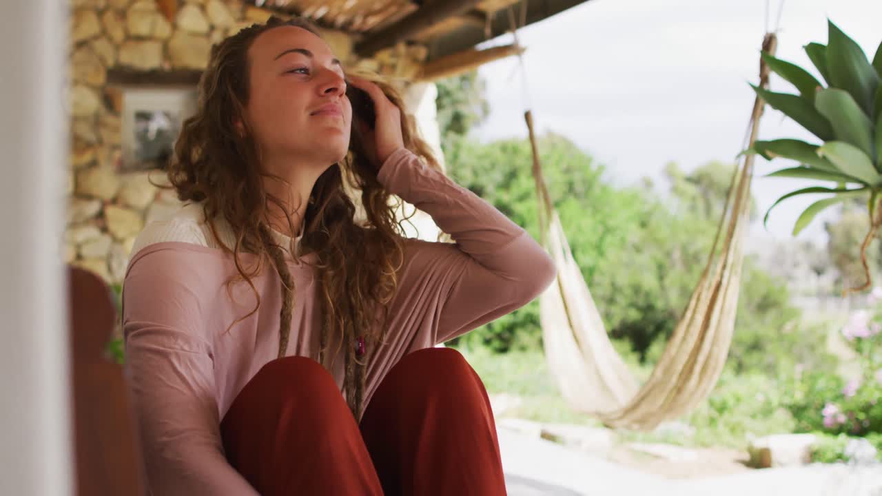mujer caucásica sonriente sentada y relajada en la terraza de la cabaña soleada, mirando al jardín