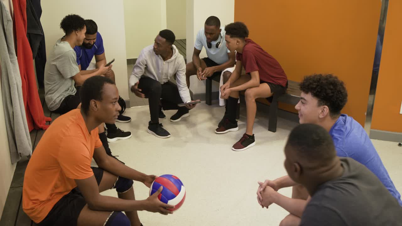 Diverses Männerteam sitzt auf Bänken in der Umkleidekabine, bespricht sich mit dem knienden Trainer, der ein Telefon mit Volleyball zeigt.
