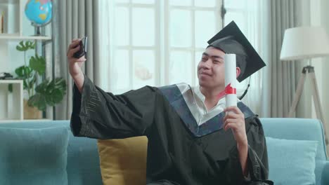 young asian man taking a selfie while wearing a graduation gown and cap, hand holding university certificate in living room