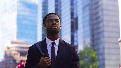 young businessman wearing wireless earbuds streaming music or podcast from mobile phone walking to work in offices in the financial district of the city of london uk