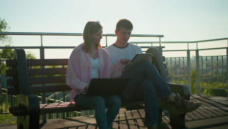 lady in pink pauses typing to show something to her seated friend who nods in agreement, both are on a bench with a backdrop of iron railings and cityscape