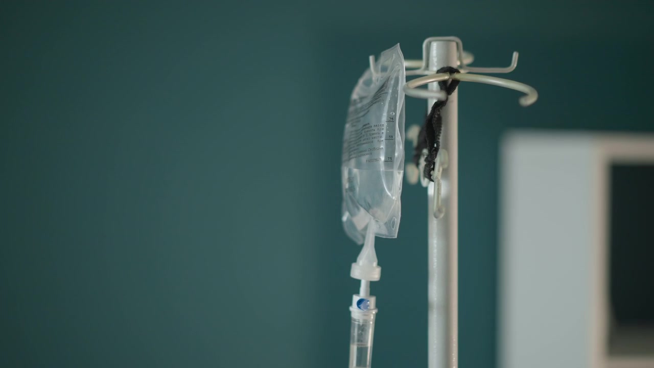Sacca sterile per flebo su asta. Sacca endovenosa ospedaliera fissata a un'asta su sfondo di parete verde acqua. Sacca sterile di liquido endovenoso medico sospesa su supporto metallico in ambiente clinico.