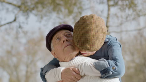 white haired senior woman embracing and kissing her husband from behind while sitting on bench in city park