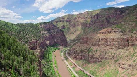 Beautiful-Large-Deep-Rocky-Canyon-Covered-In-Bright-Healthy-Foliage-And-River-Flowing-At-The-Base-With-Clear-Blue-Cloudy-Sky