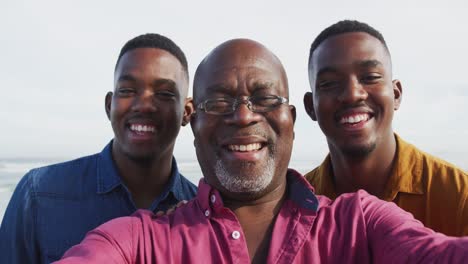 Smiling-african-american-senior-father-and-twin-teenage-sons-standing-on-a-beach-taking-a-selfie