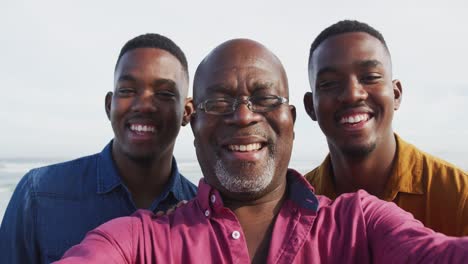 smiling african american senior father and twin teenage sons standing on a beach taking a selfie