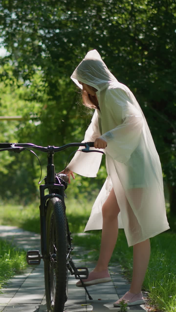 반투명한 레인코트를 입은 젊은 여성은 자전거를 타고 빛의 길에 서서 손잡이에 팔을 고 멀리서 사려 깊게 쳐다보고 있습니다. 배경에는 울창한 초록색과 나무가 있습니다.