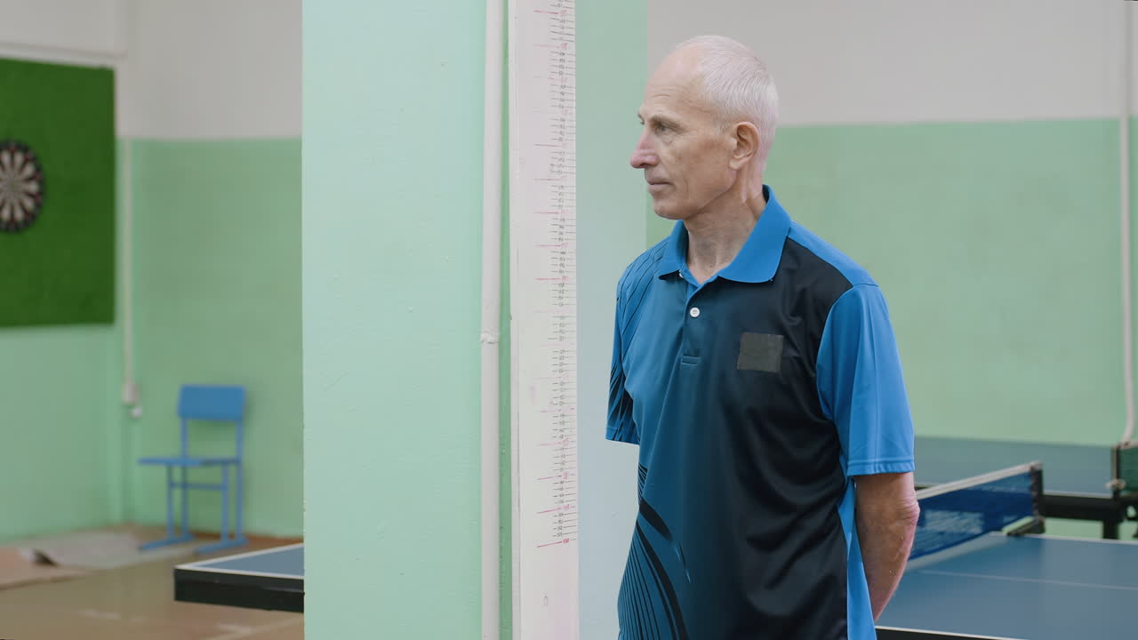 Senior coach in blue sportswear carefully watches young athlete holding paddle during table tennis training, focusing on form, precision, and preparation in indoor sports hall for skill development