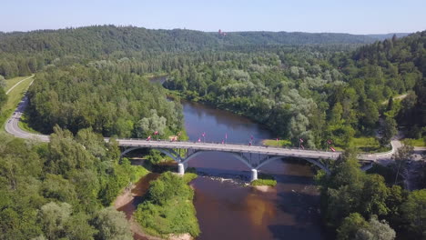 high angle aerial shot of a group of bicyclists crossing a bridge over a river in latvia