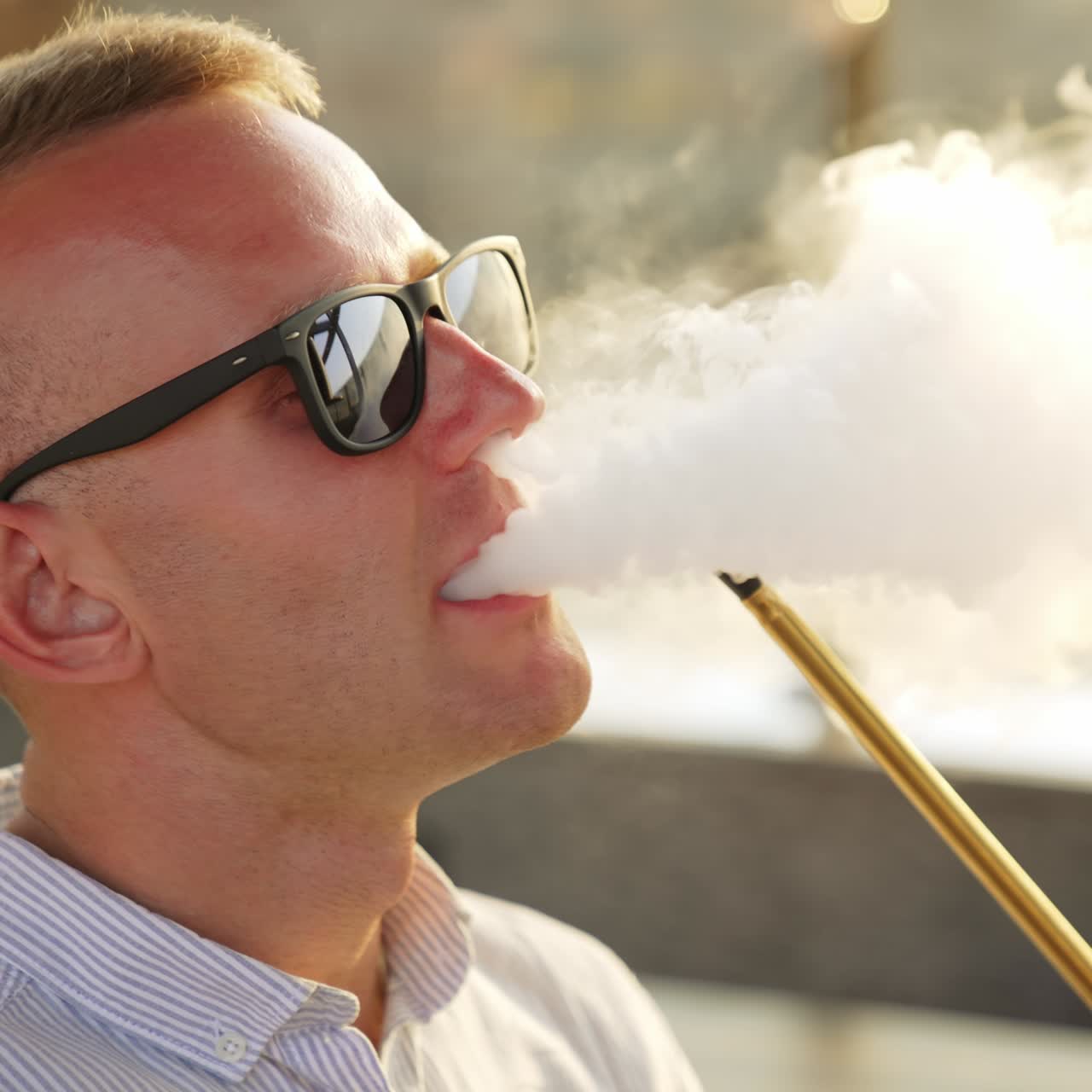 Mid-aged man in sunglasses smoking chillum sitting outdoors. Chilling male rises head and breathes out white smoke. Close up