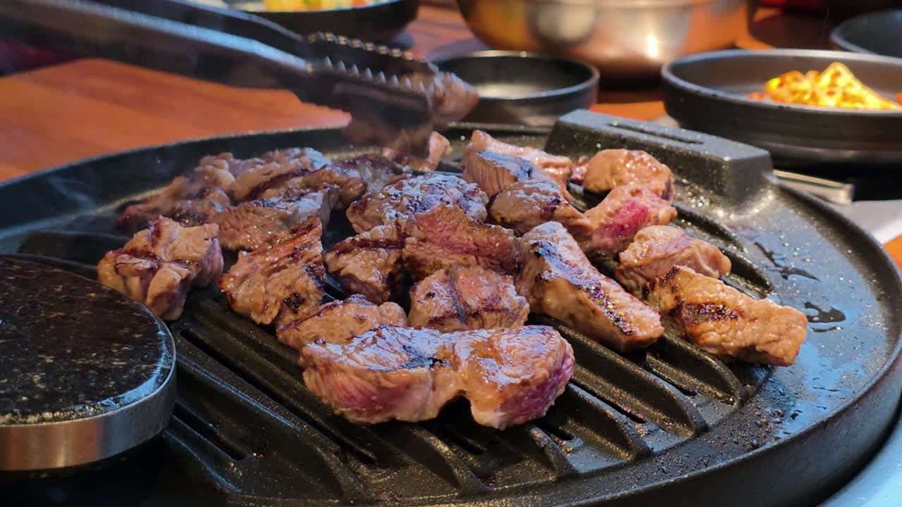 Juicy pieces of Korean BBQ beef grilling on a hot grill plate, turned with tongs, surrounded by side dishes