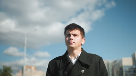 banker in black coat walking confidently outdoors with sunlight reflecting off him under bright sky in urban setting featuring blurred street light pole and buildings in background