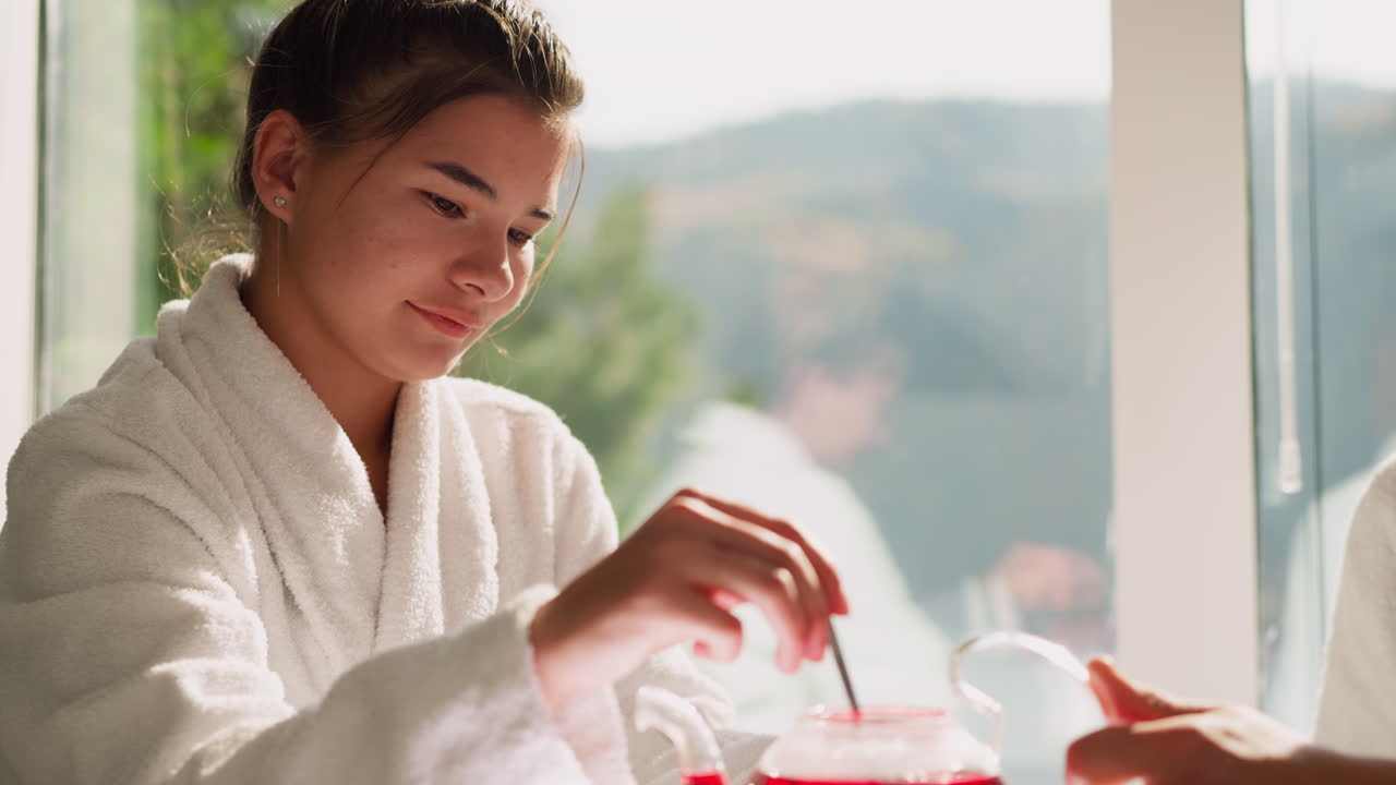 Woman stirs hibiscus tea with spoon at table closeup. Young woman in bathrobe brews hot beverage tea for husband sitting by window. Lazy morning routine