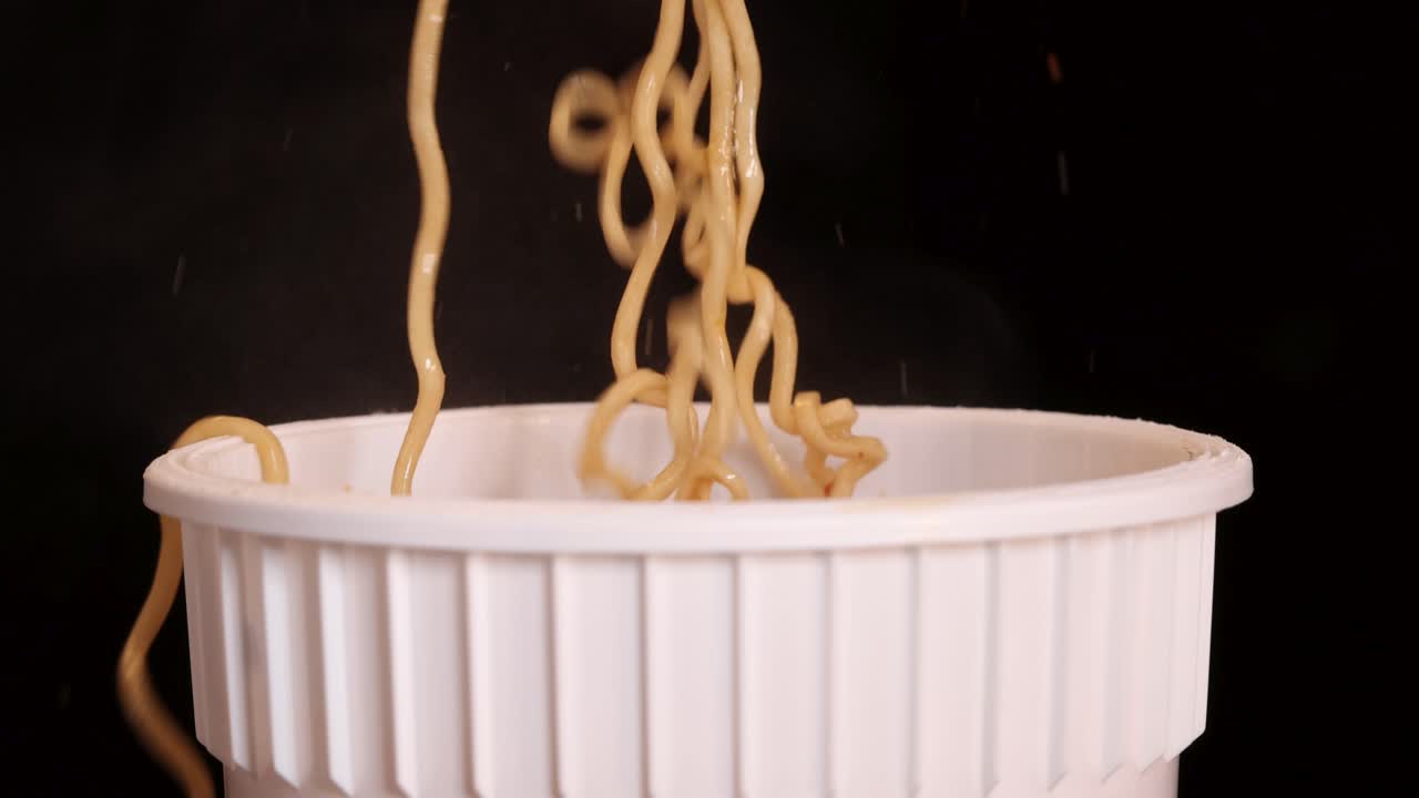 Le bacchette afferrano e sollevano noodles istantanei cotti da una tazza usa e getta bianca su uno sfondo scuro, con un'illuminazione morbida e uniforme e un movimento minimo della telecamera.