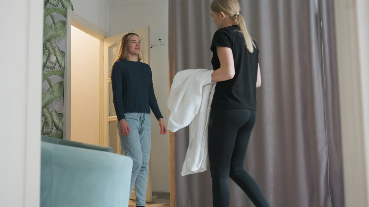 Woman walks into stylishly decorated room featuring patterned wallpaper, teal chair, and curtain divider, warmly welcomed by another lady who hands her robe in gentle indoor interaction