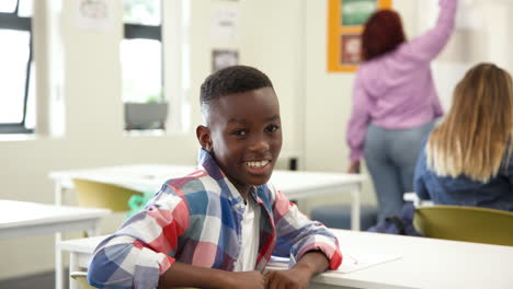 smiling african american boy in classroom looking at camera while female teacher writes, at school
