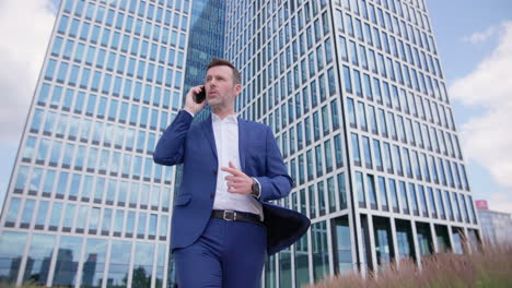 Business-man-walks-and-talks-on-phone-outside-skyscraper,-ground-view