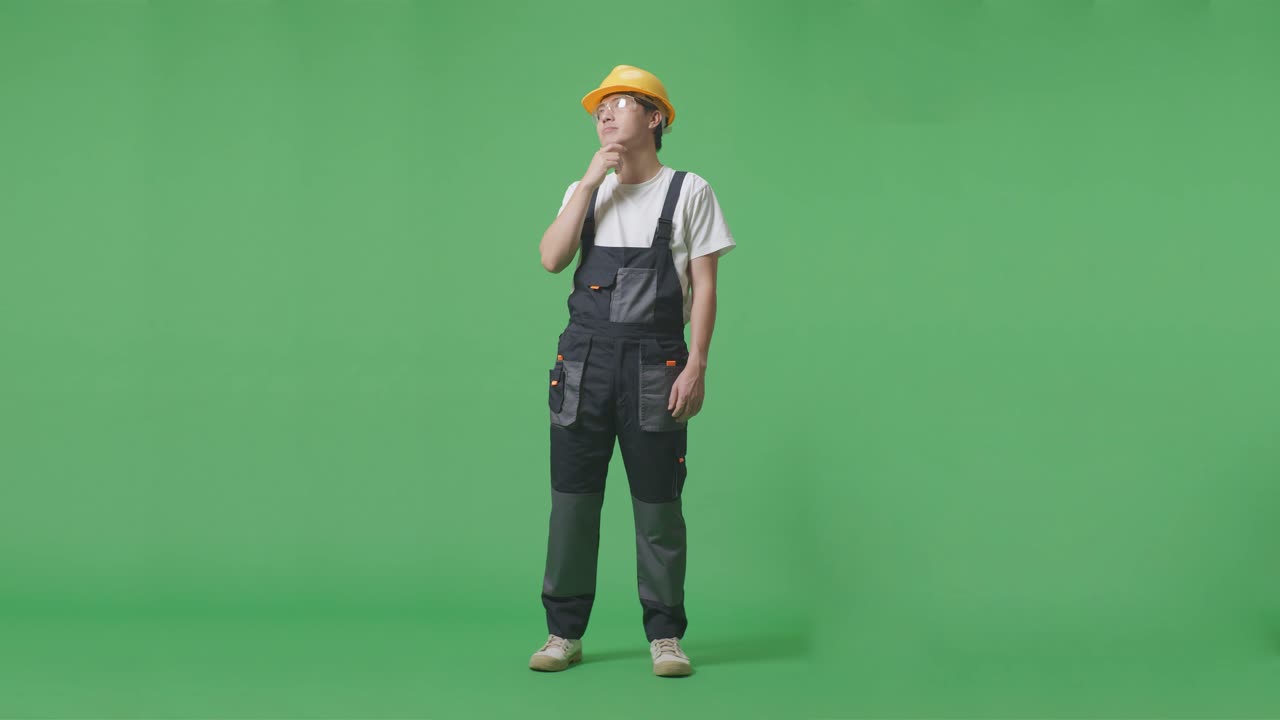 안경과 안전 헬을 착용 한 아시아 남성 노동자의 몸 전체가 녹색 스크린 배경 스튜디오에 서있는 동안 무언가에 대해 생각하고 주위를 둘러보고 있습니다.