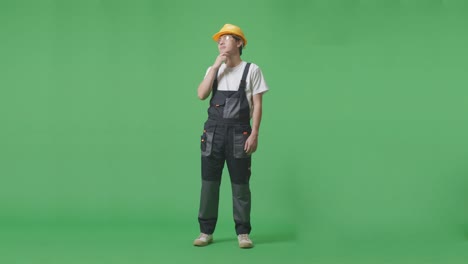 full body of asian man worker wearing goggles and safety helmet thinking about something and looking around while standing in the green screen background studio