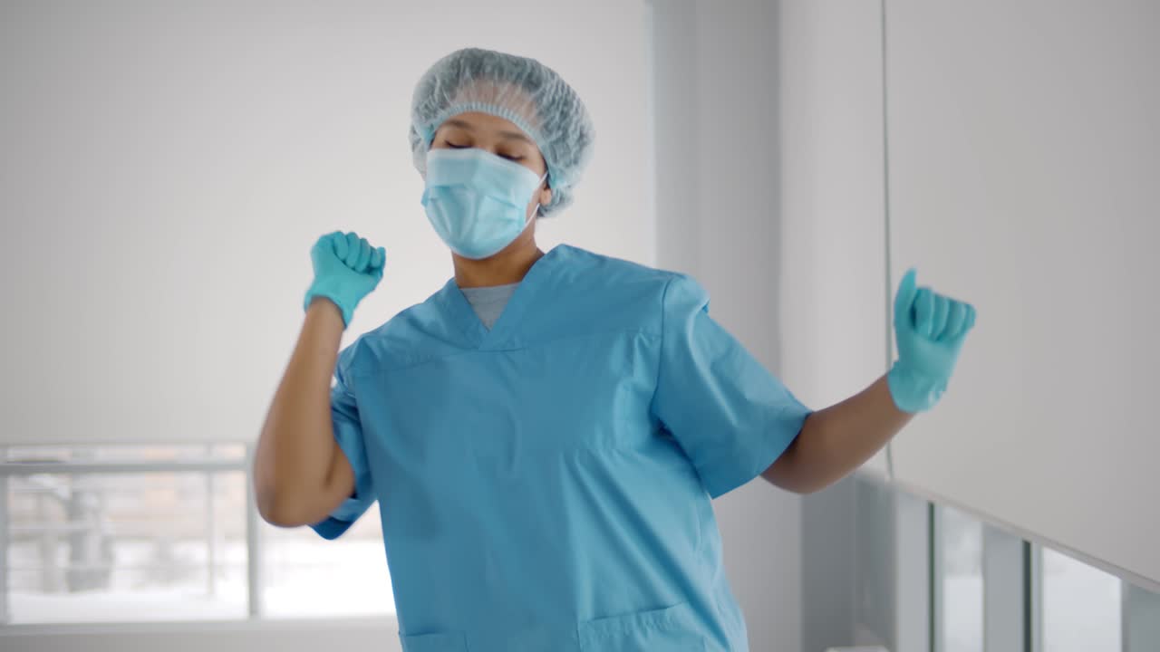 Young african american woman wearing scrubs uniform, gloves and mask dancing in hallway