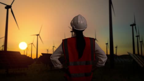 back view of female engineer in a helmet looking around while standing with arms akimbo in front of wind turbines rotating at sunset