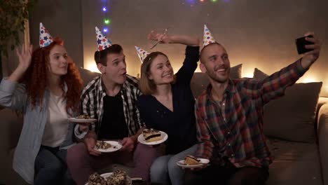 cheerful, young friends or two couples having small, cozy birthday celebration, sitting together in festive cones on the couch and taking selfie using a mobile phone 1