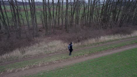 a man in a black coat and hat walking with a backpack along a row of trees along a path near a field in spring