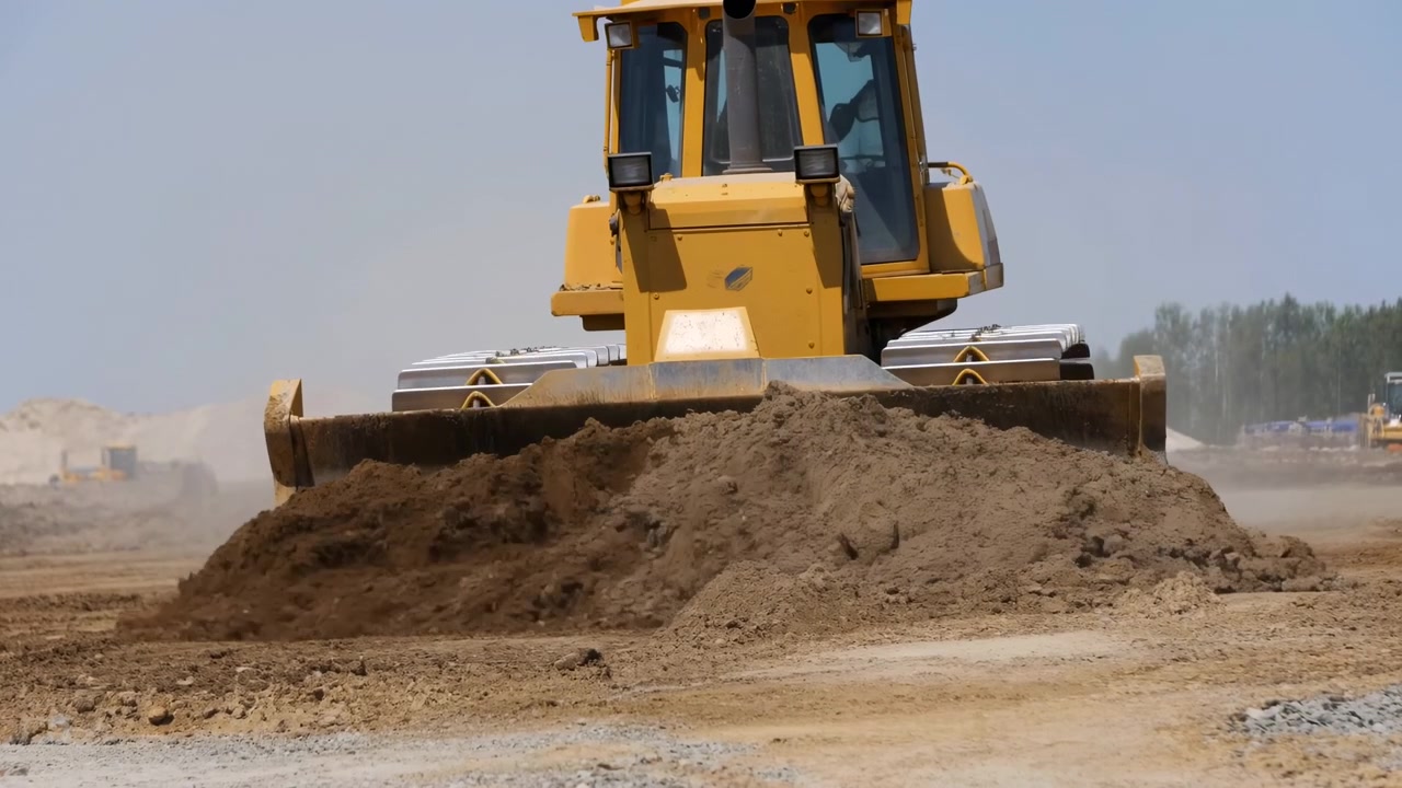 Frontalvisning viser en gul bæltebulldozer, der roligt flytter en bunke brun jord med sit brede skær, mens støv driver hen over det åbne arbejdsområde under en lyseblå himmel