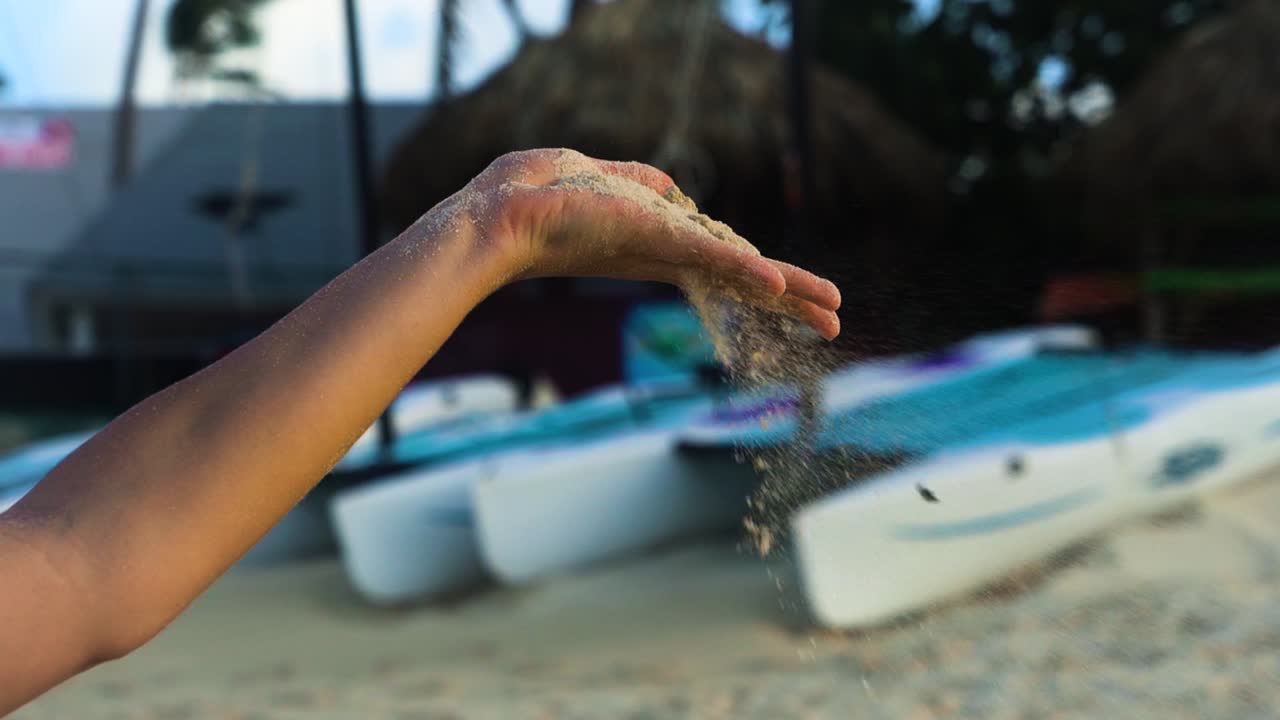 Crystal caribbean sand pours away through the woman fingers at the carribean beach with blue boats on the background