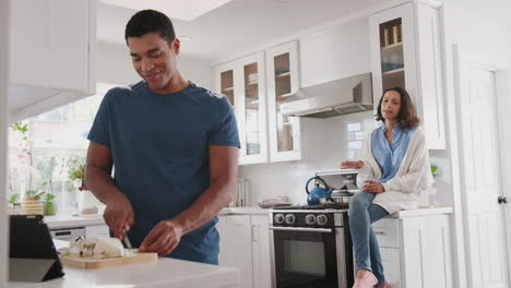 Un-Joven-Afroamericano-Preparando-Comida-En-La-Cocina,-Su-Compañero-Sentado-En-La-Encimera-De-La-Cocina-Detrás-De-él