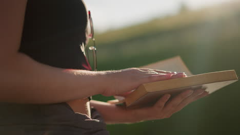 sunlight shines from blurred greenery in the background, reflecting off book held by woman wearing glasses on her black top, she gently flips through pages while seated