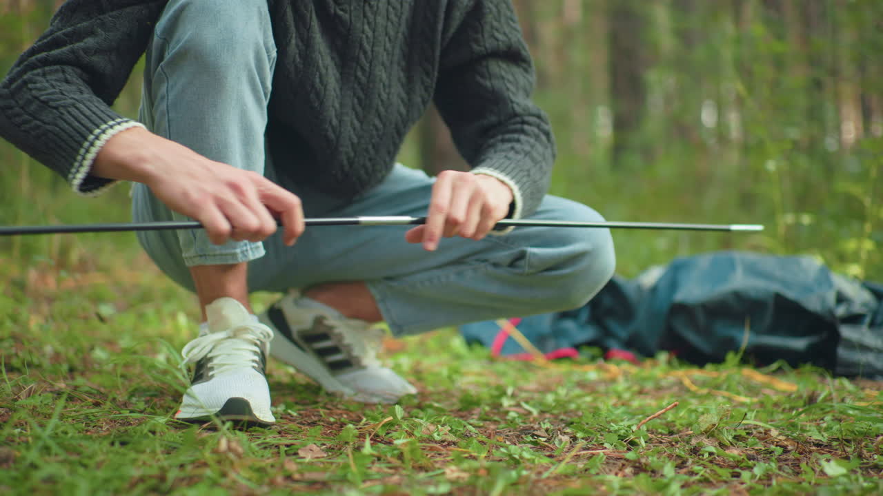 Close up of person handing tent poles to man squatting in forest as he joins them together for camping setup amid sunlit green foliage and scattered camping gear and fabric pieces outdoors