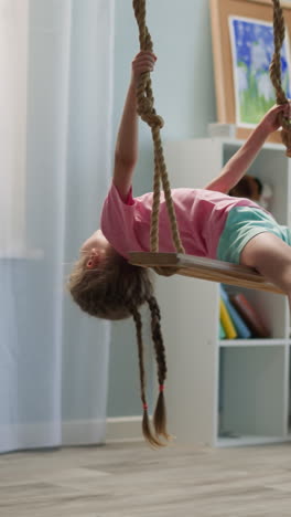 una niña rubia de preescolar con trenzas largas se sienta en un columpio en una habitación ligera con un estante de madera. un niño alegre inclina la cabeza hacia atrás para bromear sosteniendo cuerdas