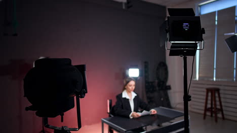 positive smiling lady wearing white shirt and black jacket sits at desk in photo studio. female reporter id getting ready for the footage in light of soffits.