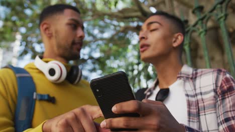 dos felices amigos varones de raza mixta de pie, hablando y usando un teléfono inteligente en la calle