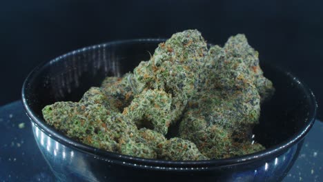 green dried marijuana buds close up concept shot, pile of dried marijuana plants, orange trichomes strains, in small black shiny bawl, on a rotating stand, studio lights, slow motion, 4k video, smoke