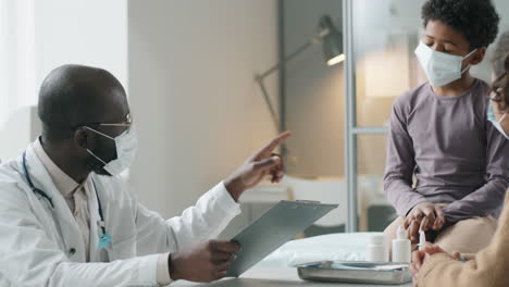 Black-Pediatrician-in-Mask-Speaking-with-Boy-and-His-Mother-in-Medical-Office