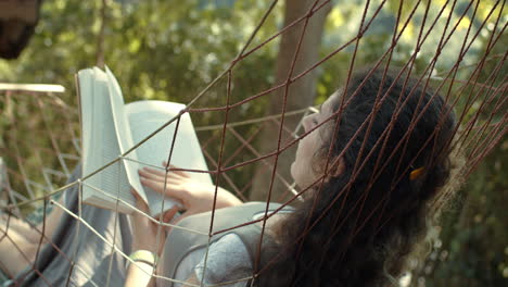 mujer consciente balanceándose en una hamaca y leyendo un libro mientras el viento sopla su cabello en cámara lenta