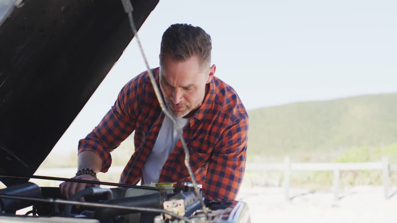 Stressed caucasian man looking at broken down car with open bonnet on sunny day at the beach