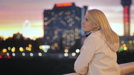 a pensive woman stands on a bridge in the night city against the backdrop of bright lights and looks