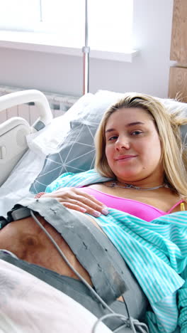 blonde pregnant woman lies in bed with cardiotocography belt on the belly. lady smiles to the camera. top view. vertical video