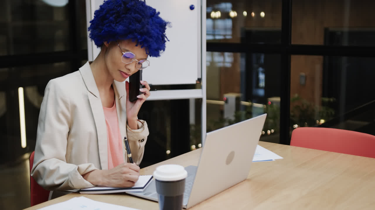 Biracial businesswoman with blue afro talking on smartphone and using laptop at desk, slow motion