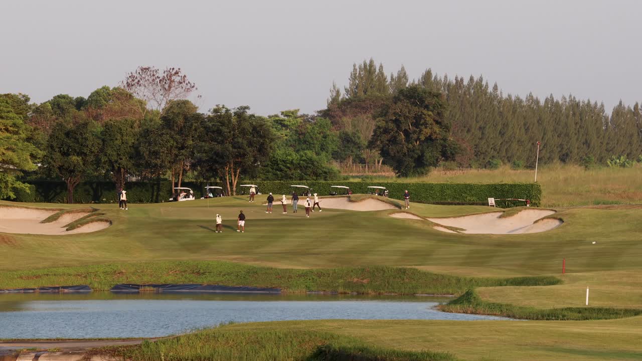 Golfers traverse lush fairway near lake, sand traps, and trees, with warm sunset lighting
