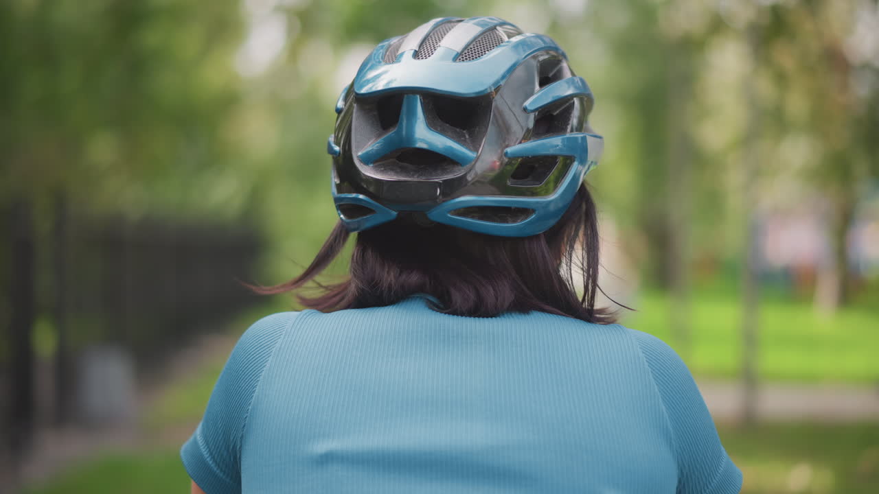 woman in helmet riding through park, back view of female cyclist on treelined pathway, urban commute mood with calm reflective atmosphere, soft green bokeh, blue top, safety focus, gentle motion